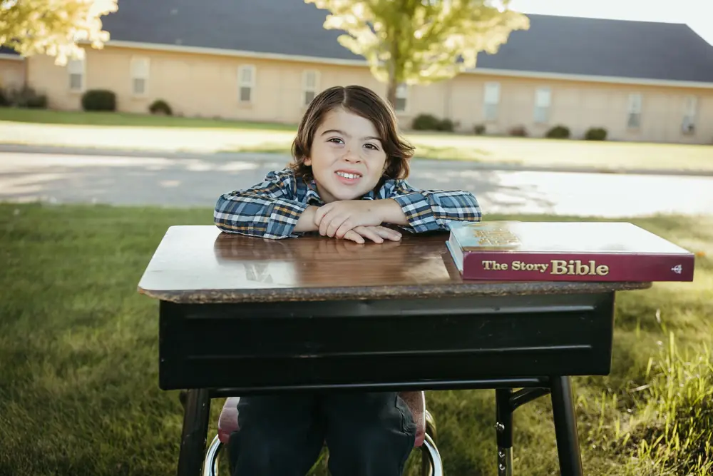 A Cornerstone Community Schoolhouse student at a wooden school desk on the lawn with The Story Bible, Caldwell, Idaho